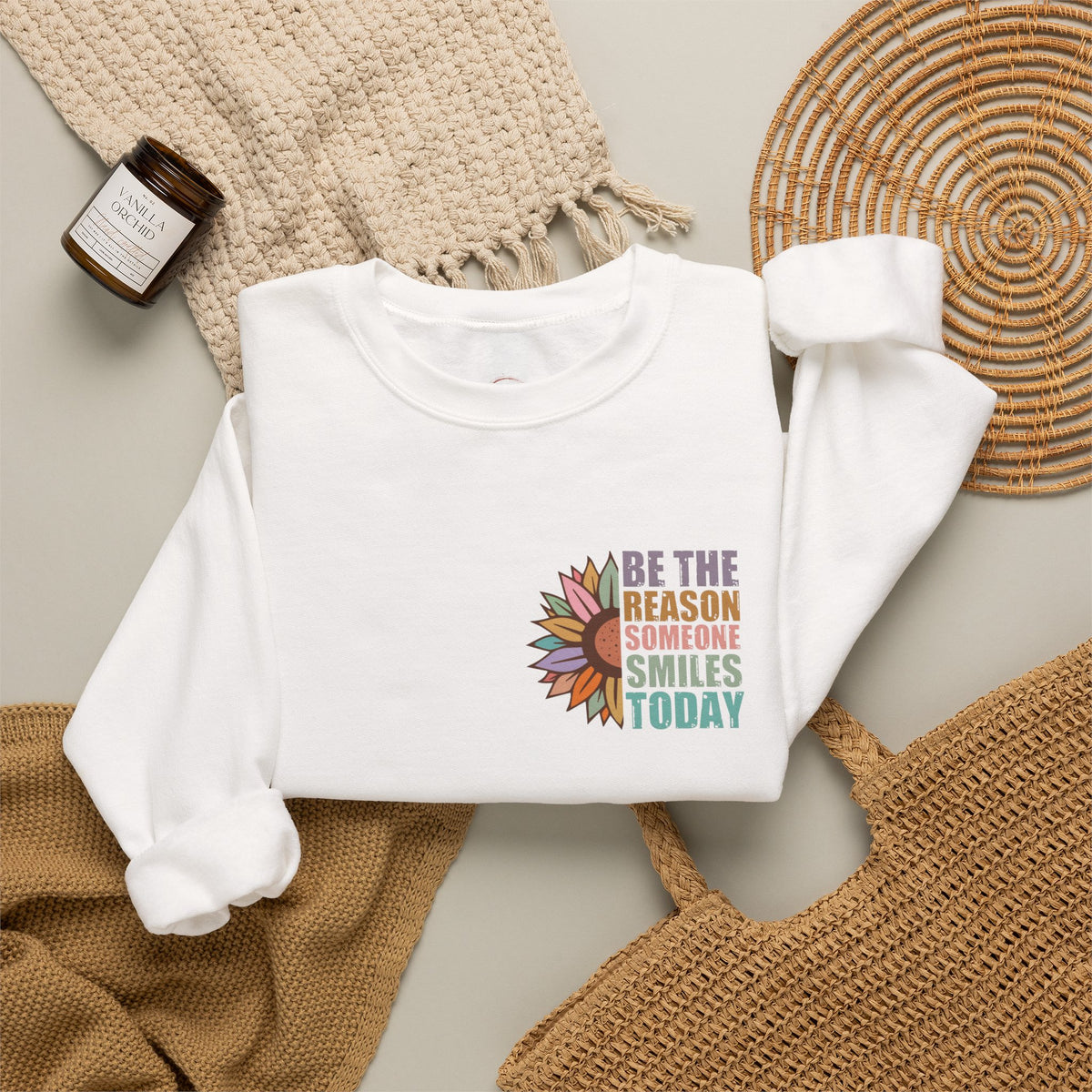 A white long-sleeve crewneck sweatshirt laid flat on a neutral background, styled with a beige knit blanket, a woven straw hat, a small candle jar, and a tan woven mat. The sweatshirt features a colorful graphic of a sunflower and the text 'BE THE REASON SOMEONE SMILES TODAY'.