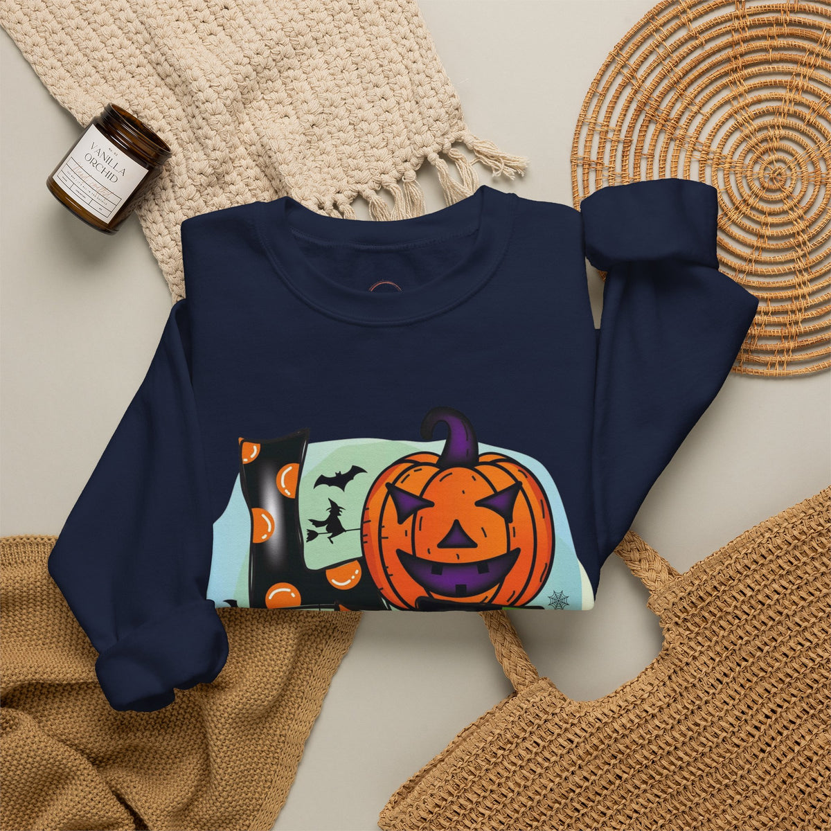 A navy blue sweatshirt with a colorful Halloween graphic featuring a jack-o'-lantern, bats, and a witch hat, styled in a flat lay with a knit blanket, woven placemat, and a candle.