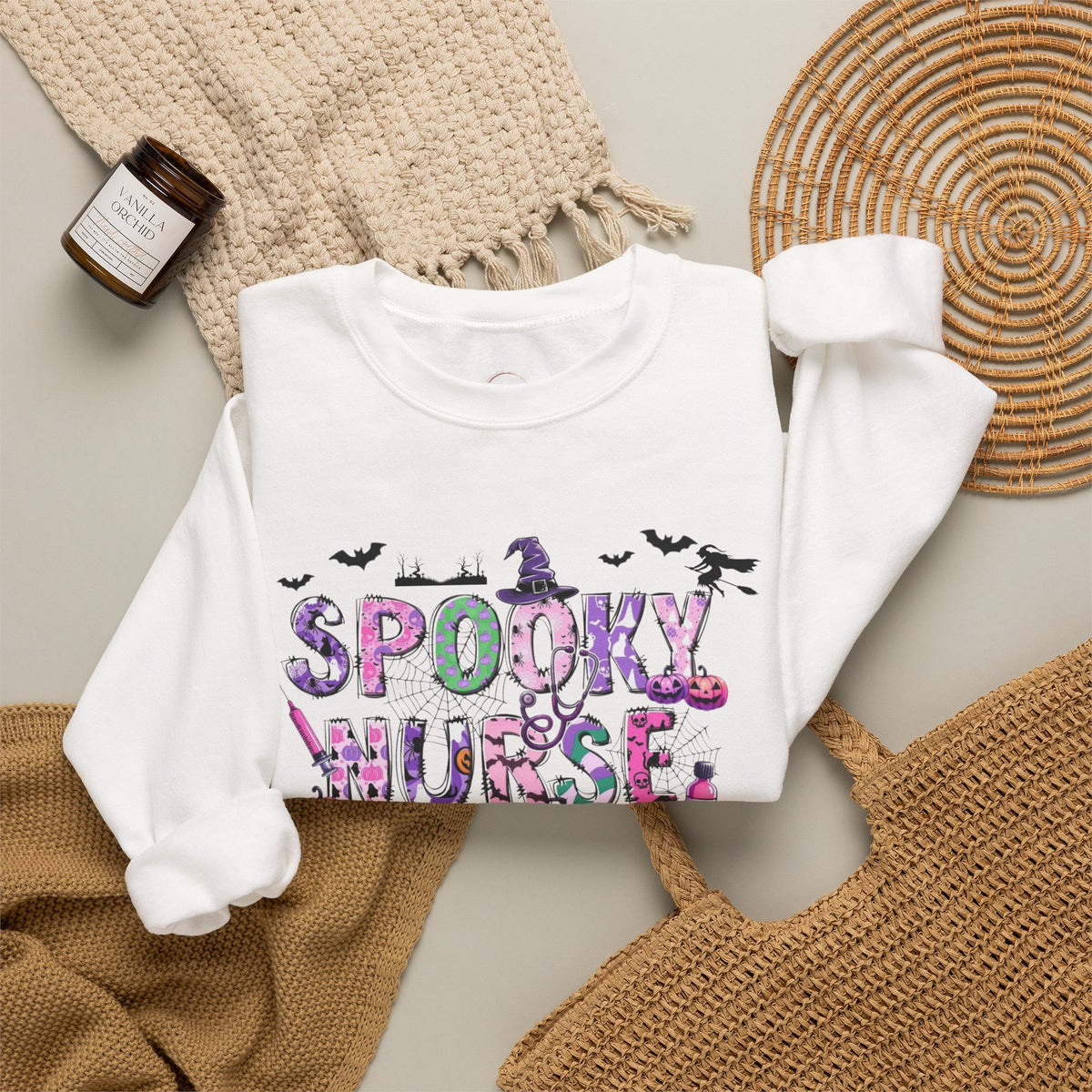 A white long-sleeve sweatshirt with a colorful 'Spooky Nurse' graphic, styled in a flat lay with a knit blanket, a woven placemat, a small candle, and a brown woven bag on a neutral background.
