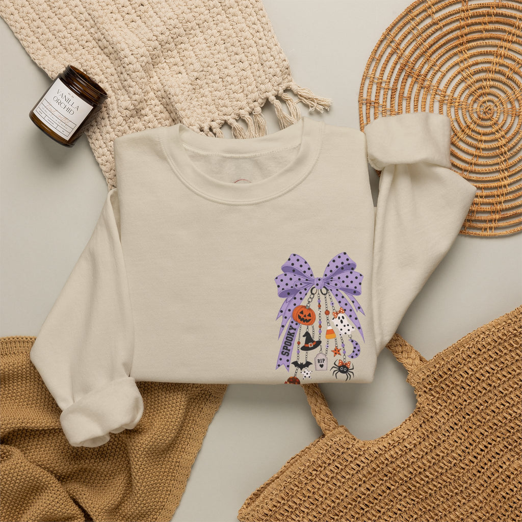 An off-white long-sleeve crewneck sweatshirt with a Halloween-themed graphic print featuring a purple polka-dot bow, a jack-o'-lantern, a witch, and a spider. The sweatshirt is styled in a flat-lay arrangement with a beige knit blanket, a woven basket, a small candle, and a wicker placemat.
