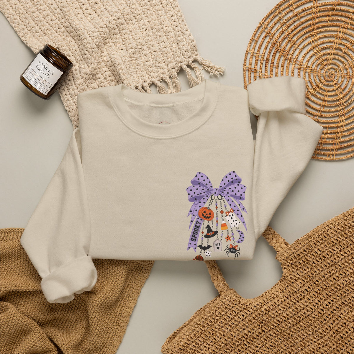 An off-white long-sleeve crewneck sweatshirt with a Halloween-themed graphic print featuring a purple polka-dot bow, a jack-o'-lantern, a witch, and a spider. The sweatshirt is styled in a flat-lay arrangement with a beige knit blanket, a woven basket, a small candle, and a wicker placemat.